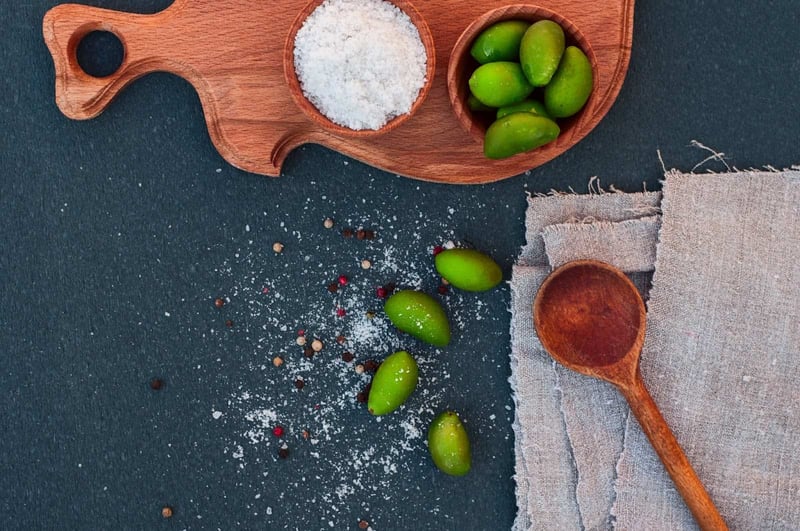 Delicious green olives, salt, and peppercorns arranged on a dark textured surface for cooking or snacking.