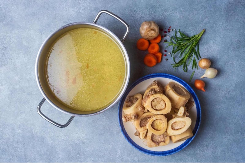 Healthy dog bone broth with marrow bones, garlic, carrots, and fresh herbs on a blue surface.