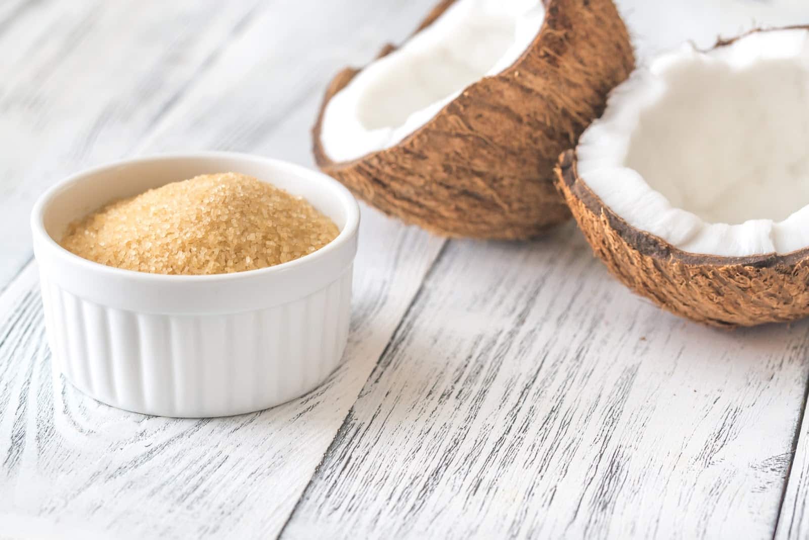 Natural coconut sugar in a white ramekin with split coconut shells on rustic wooden surface.