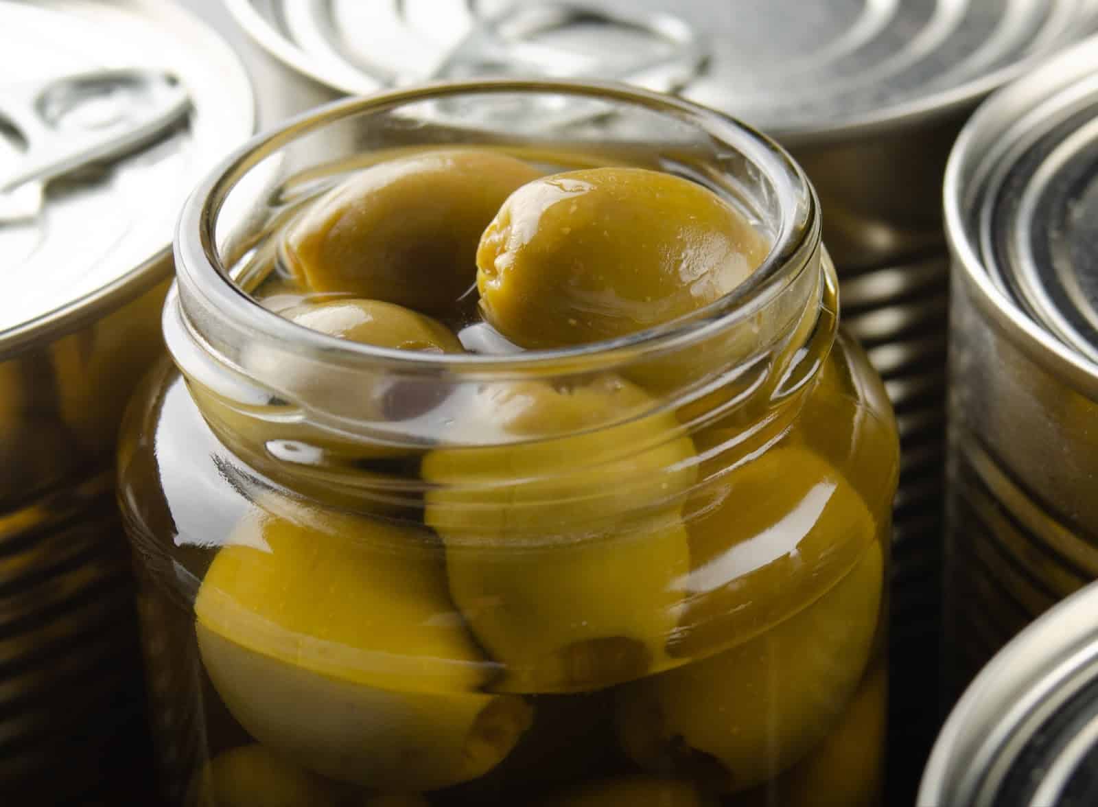 Delicious green olives in a glass jar, perfect for snacking or cooking, on a background of unopened canned goods.