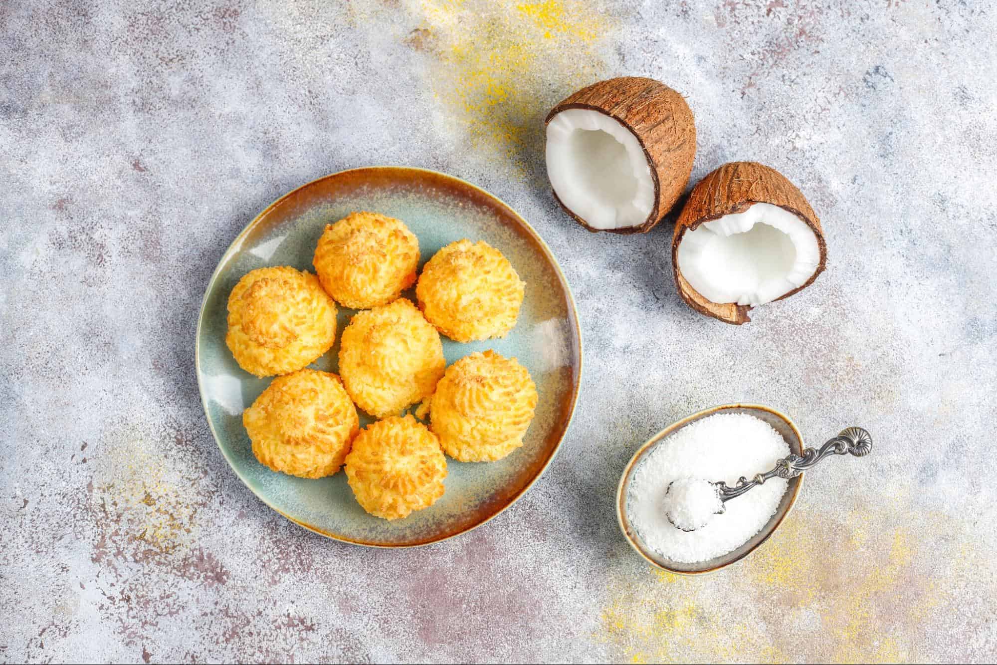 Golden coconut macaroons on a teal plate, with fresh coconut halves and shredded coconut on a textured background.