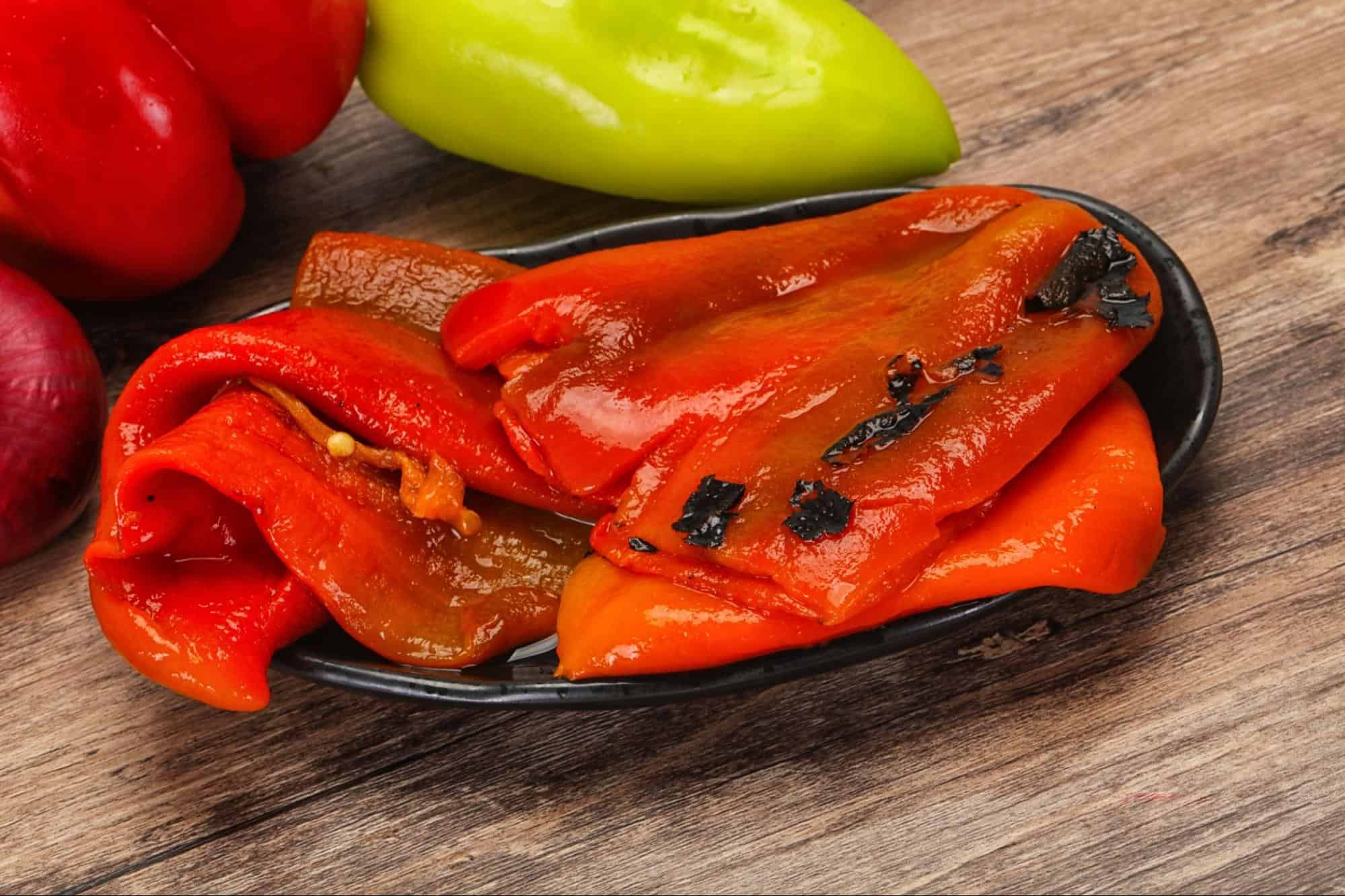 Close-up of charred red bell peppers on a black dish with fresh peppers in the background.