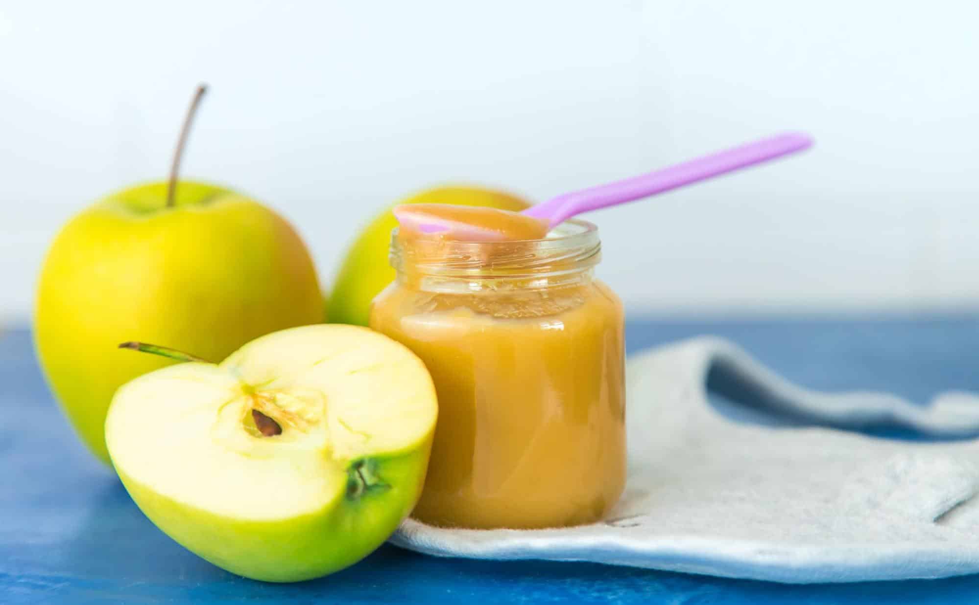 ALT text: Fresh apples and dog-friendly apple puree in jar with a spoon on a blue surface.