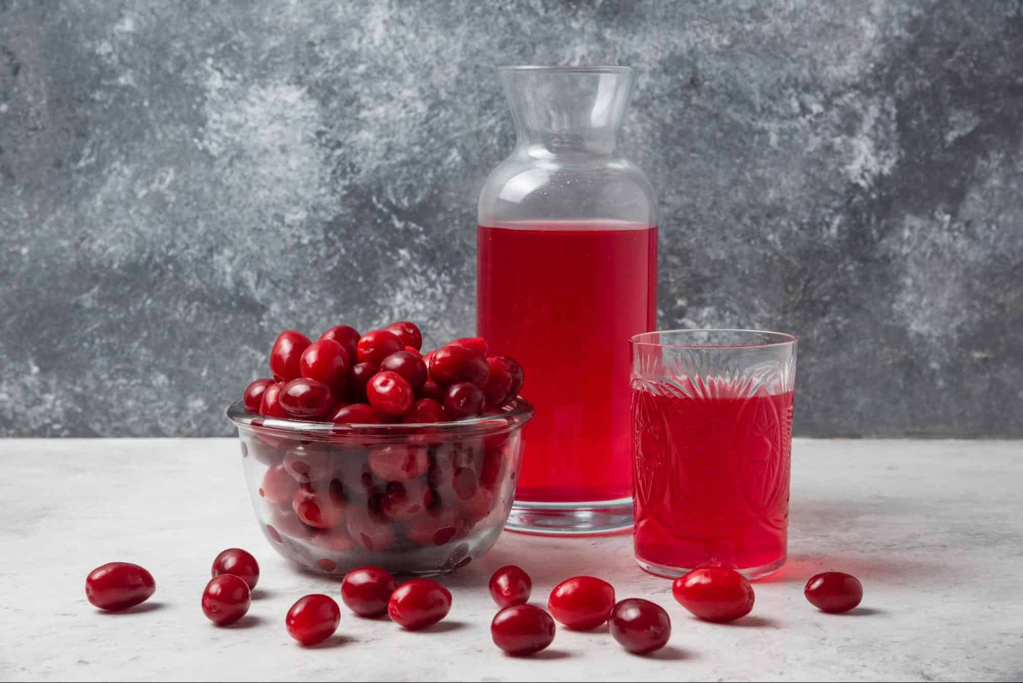 Rich cherry juice in a glass pitcher and a glass of cherry juice, alongside fresh cherries, on a white surface with a textured grey background.
