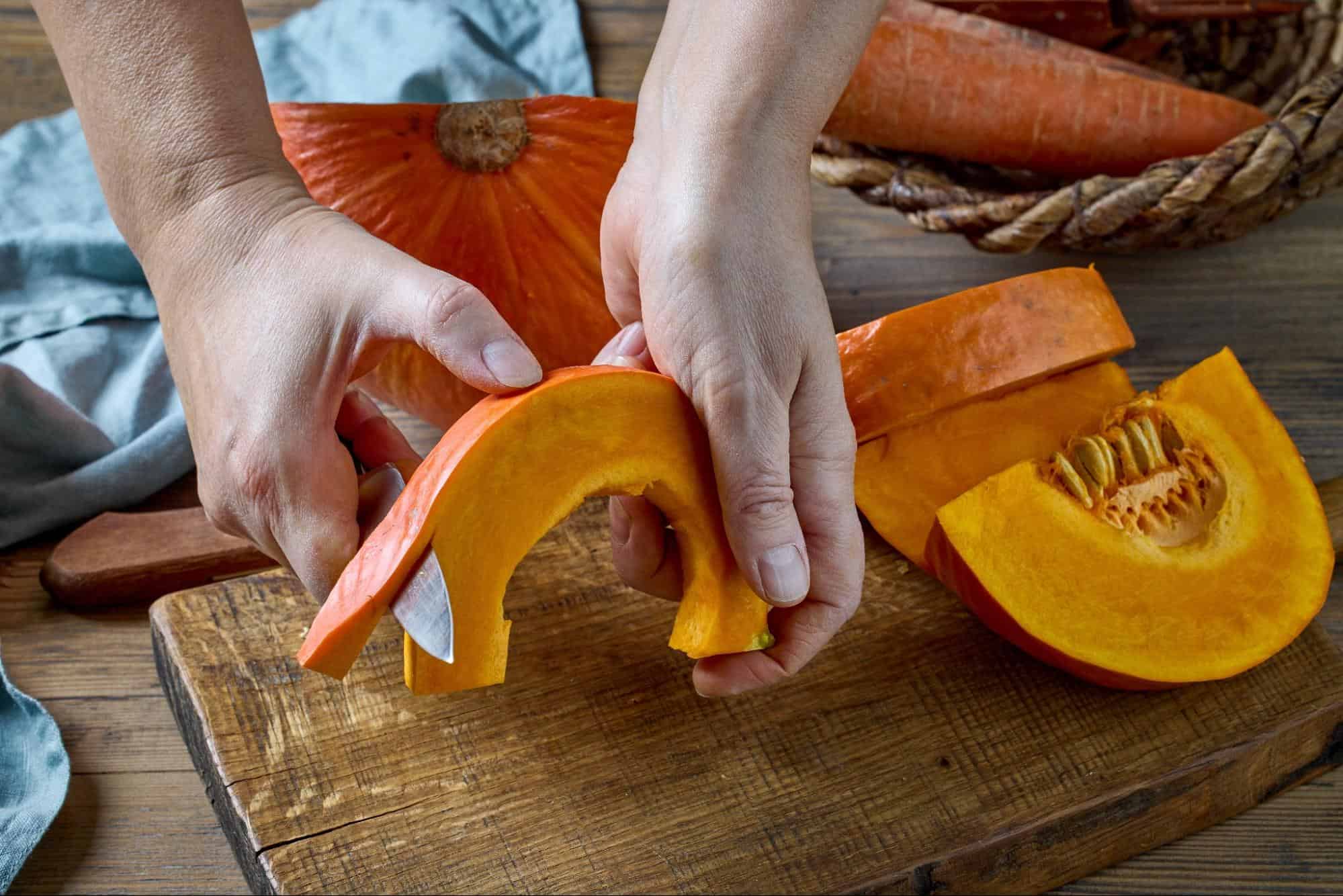 Close-up of hands slicing fresh orange pumpkin on wooden cutting board.