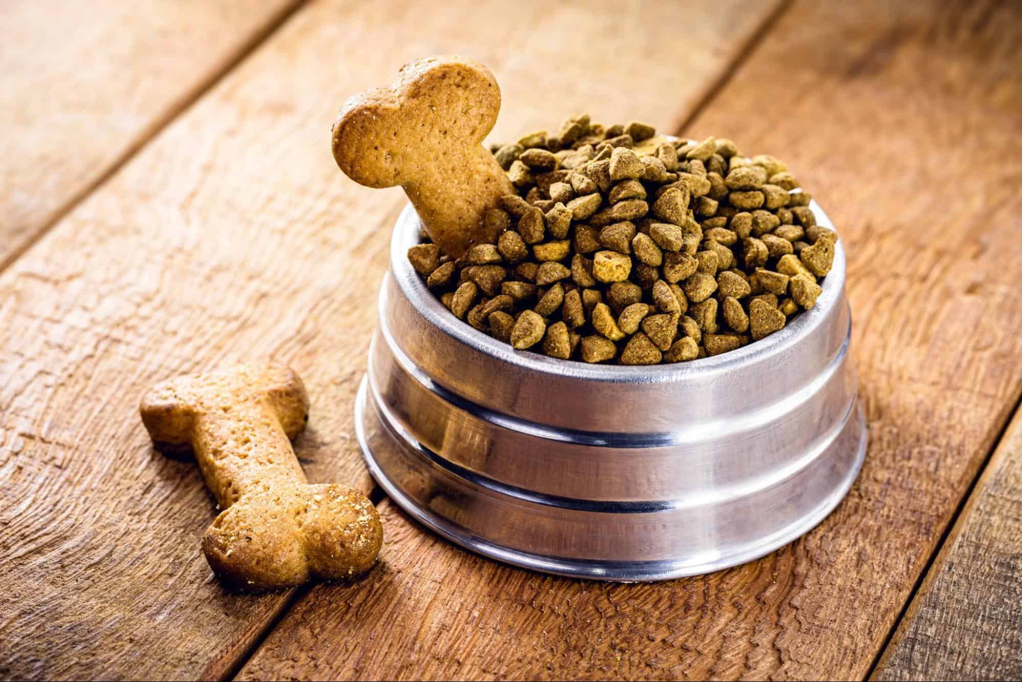 Dog food in stainless steel bowl with bone-shaped dog treats on wooden surface.