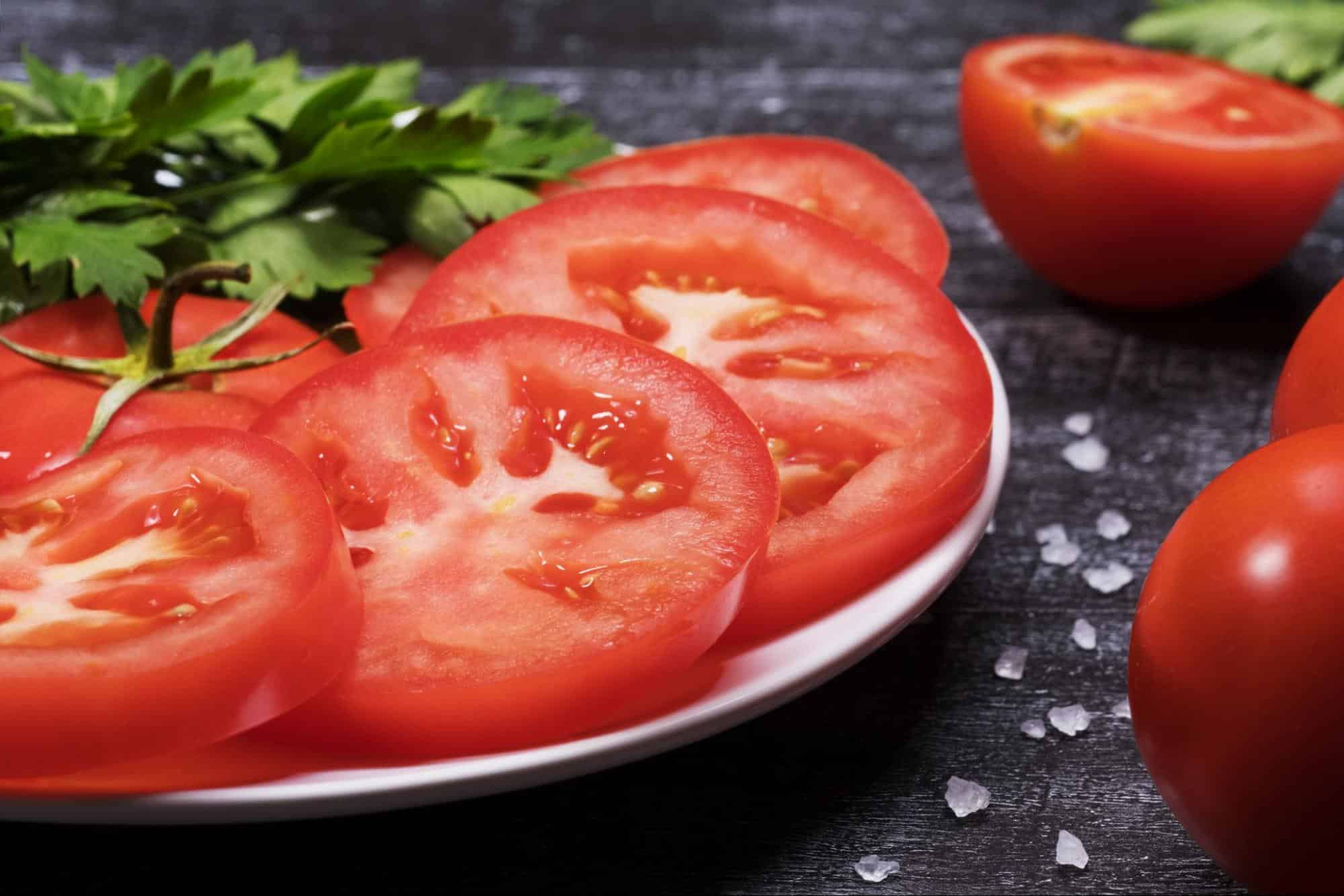 Bright, fresh tomatoes sliced for salads or cooking, on dark rustic wood with herbs and salt.