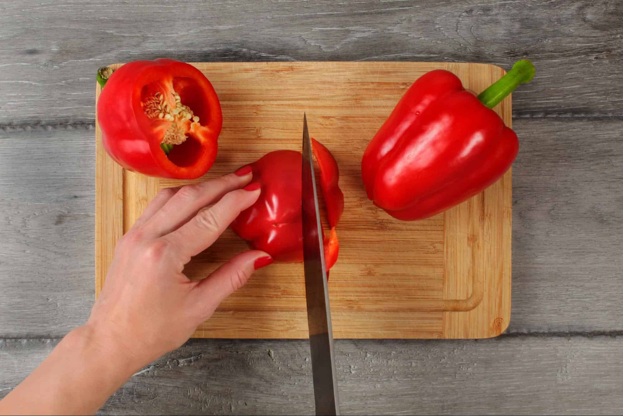 Close-up of a hand slicing a red bell pepper on a wooden cutting board for healthy dog treats.