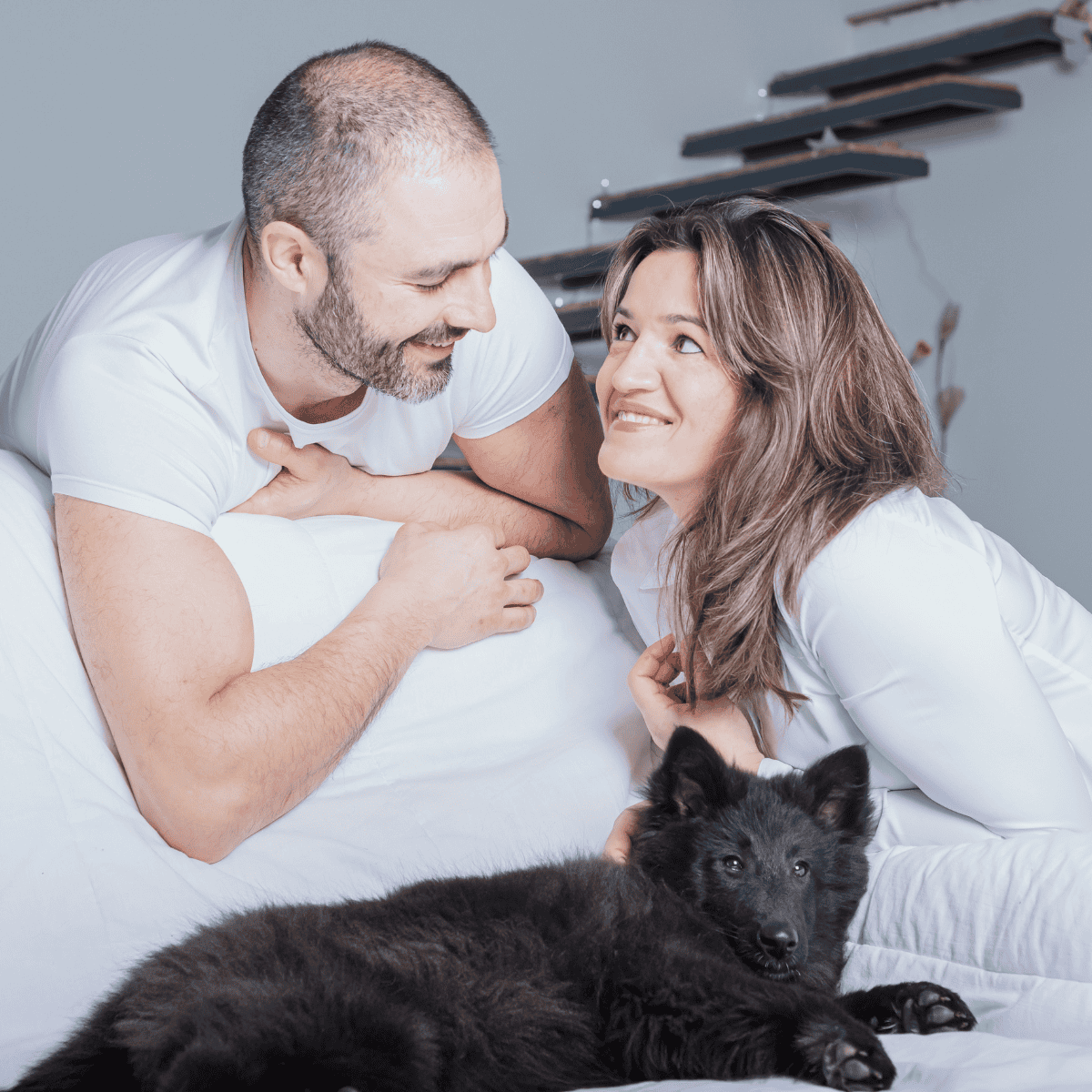 Close-up of a joyful couple with their black puppy lying on a bed, showcasing pet care and companionship.