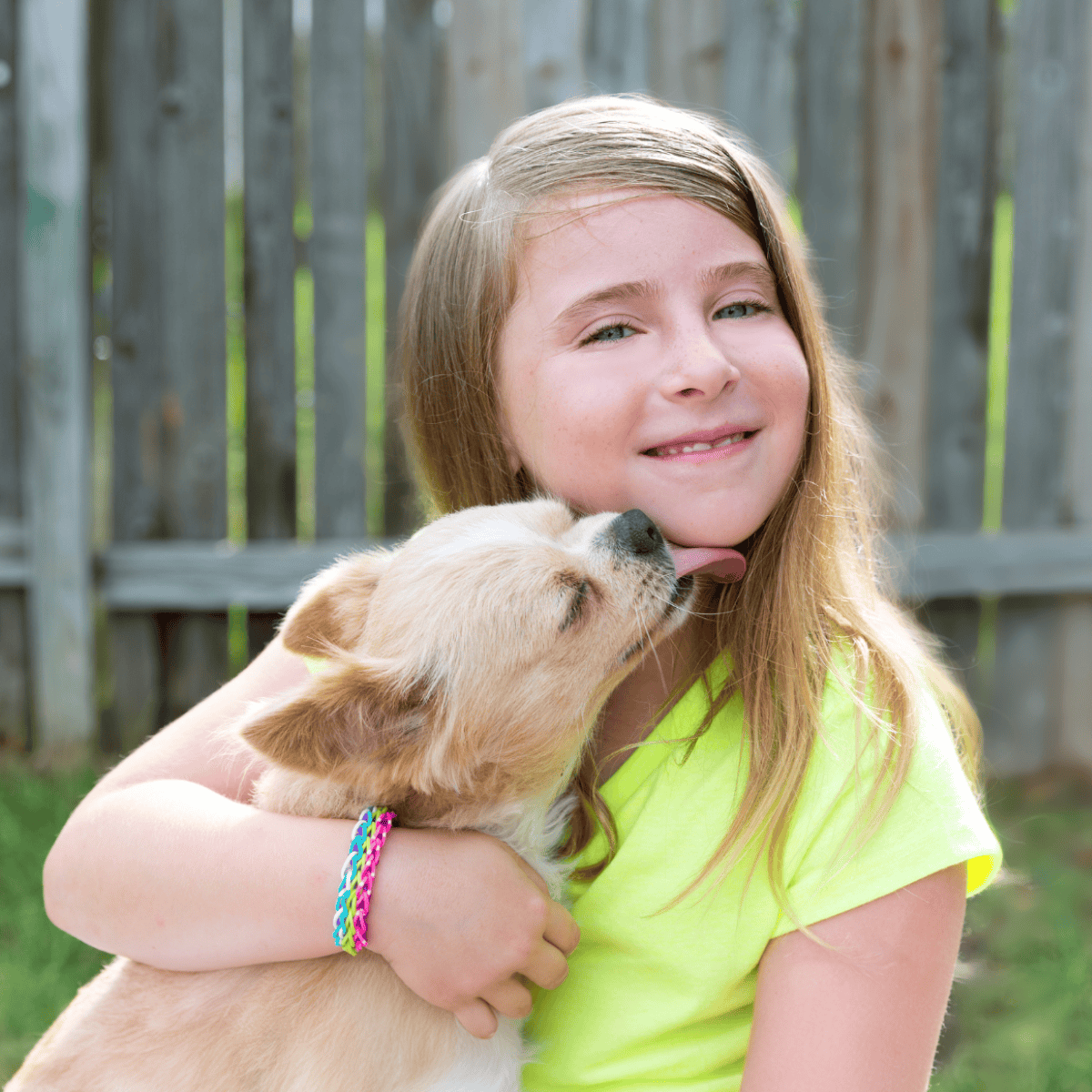 Adorable girl hugging puppy outdoors, smiling with affection, wooden fence background, joyful pet bonding.
