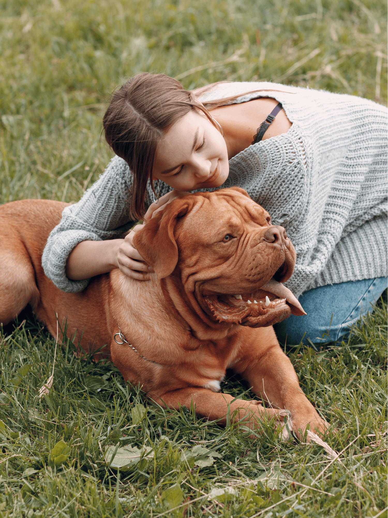 Adorable woman cuddling her large brown dog on a grassy field.