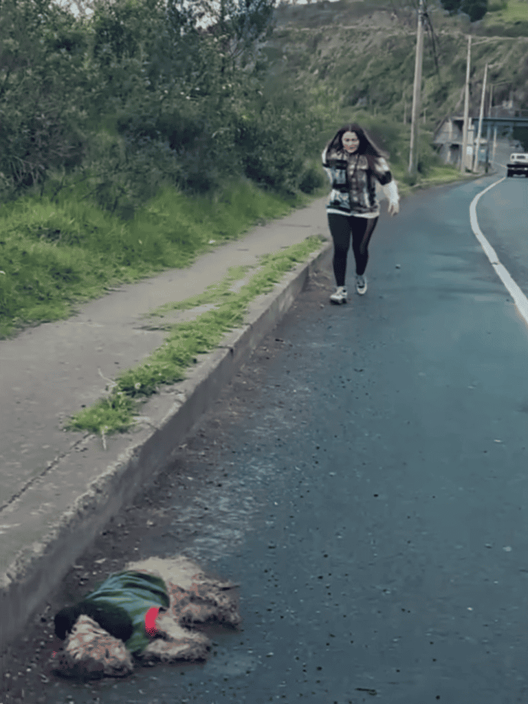 Dog relaxing on the road during walk, wearing a green jacket, with person approaching in background.