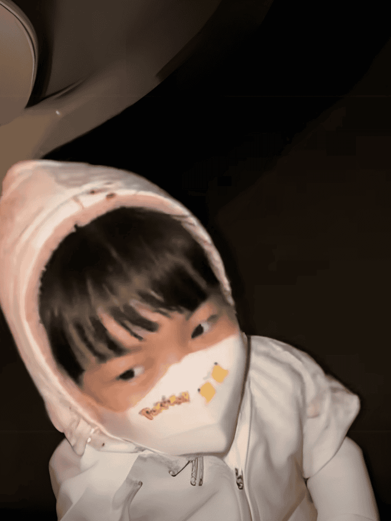 Child wearing a face mask and hoodie, looking at the camera indoors.