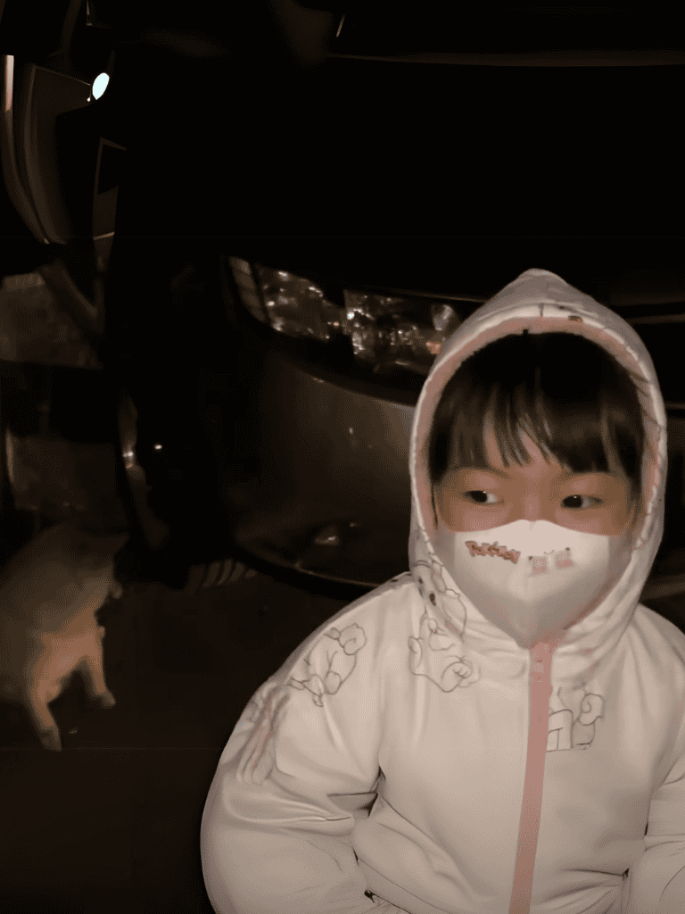 Close-up of a young girl wearing a mask standing near a dark car at night, outdoors.